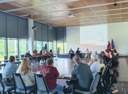 Udeleženci sedijo za mizo in poslušajo predavatelja. Na veliki beli steni je predstavitev, ker so podatki o razdeljevanju hrane.